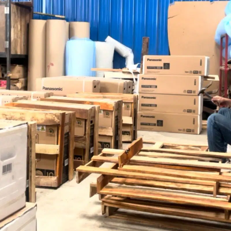 Deretan elektronik besar bermerek Panasonic dan beberapa peti kayu di area gudang sebagai ilustrasi layanan tarif kirim mesin dan elektronik besar.